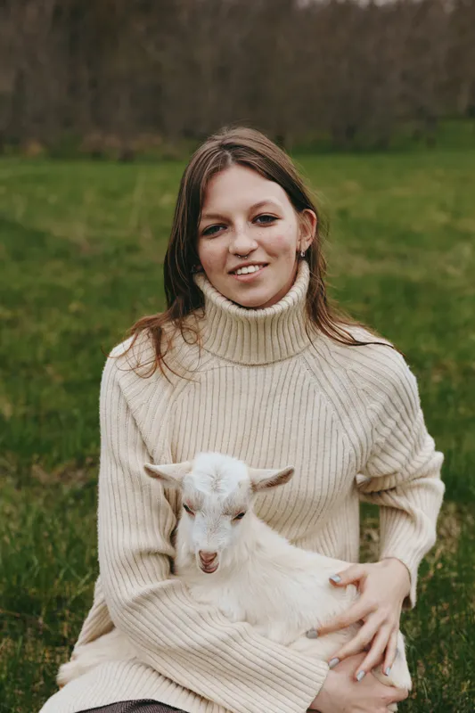 A smiling woman sits on grass in a cream turtleneck, holding a white goat kid. Bright, calm countryside setting communicates warmth, connection, and rural lifestyle.