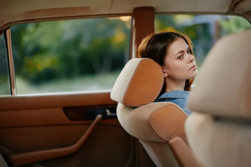 Young woman in casual denim attire sitting in a car, looking thoughtfully out the window, capturing a moment of contemplation and serenity amidst nature.