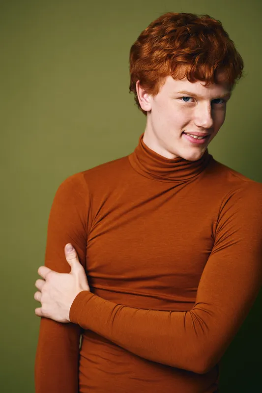 Young man with red hair in brown turtleneck sweater posing with crossed arm looking at camera against green background closeup portrait.