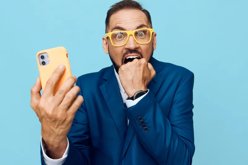 A business professional stands against a vivid blue backdrop, taking a candid selfie with a bright yellow phone, showing a moment of surprise and intrigue.