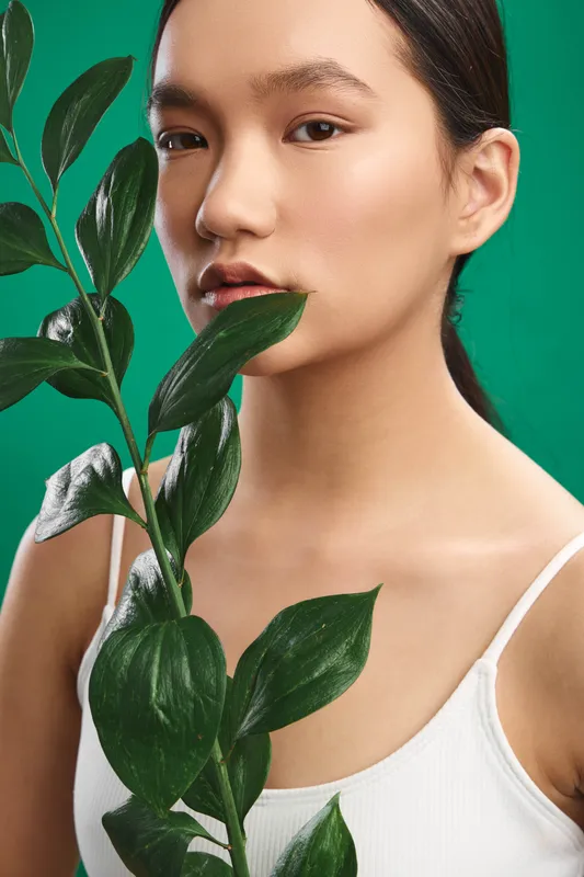 Asian woman stands with a leafy plant in front of a vibrant green backdrop, presenting a clean, natural portrait that conveys calm focus, serenity, and modern simplicity.