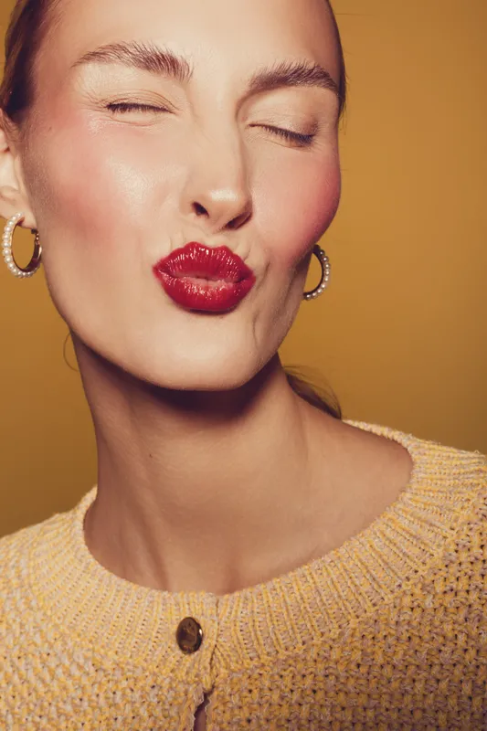 Close-up of a woman with red lipstick making a kiss face, wearing hoop earrings and a knitted sweater. Warm lighting highlights her smooth skin and playful expression on orange background.