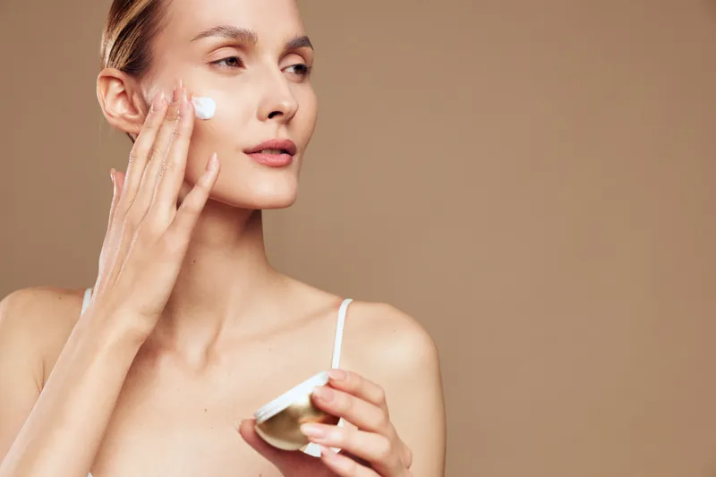 Young woman applying moisturizing cream on face with gentle touch, neutral background, natural skin care routine, soft lighting, minimal makeup, focused expression