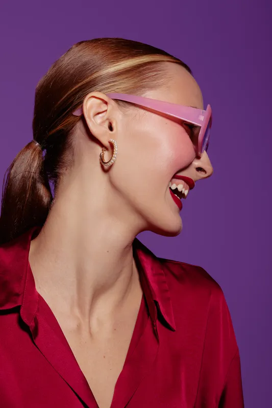 Smiling woman with red hair tied in ponytail wears pink sunglasses and red shirt. Side profile on purple background. Concept of fashion, style, happiness, and modern portrait.