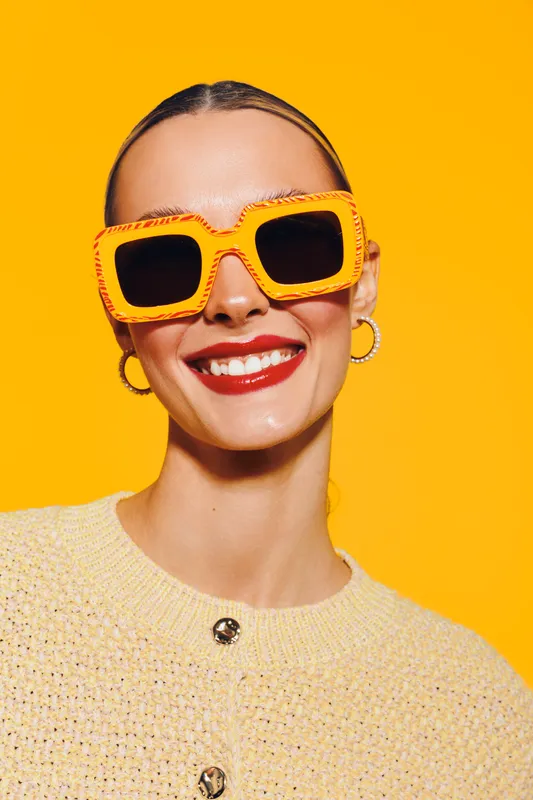 Smiling woman with bright red lipstick wearing large orange sunglasses and yellow sweater on vibrant yellow background. Concept of fashion, summer style, and confident mood.