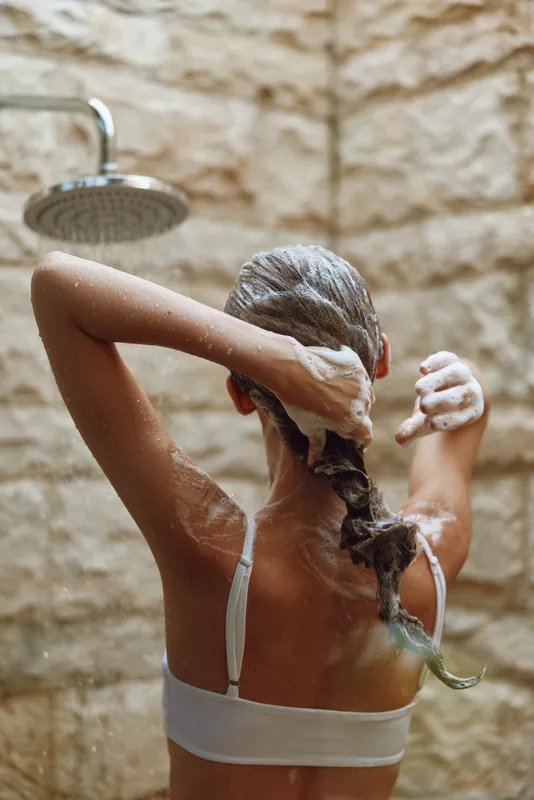 Woman washing hair in the shower with foam and water droplets, natural stone wall background, warm lighting, spa-like atmosphere, personal care moment.