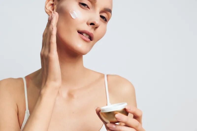 Young woman applies facial cream on clean skin holds jar of moisturizer skincare beauty routine isolated on light background with natural fresh healthy appearance.
