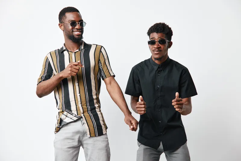 Two young black men with dark skin wearing sunglasses and casual shirts standing together on white background smiling and posing for camera with confident and relaxed mood.