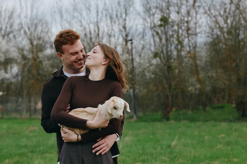 Young couple embraces outdoors as the woman holds a small goat in her arms while they smile warmly enjoying a peaceful moment in a green field with bare trees in the background.