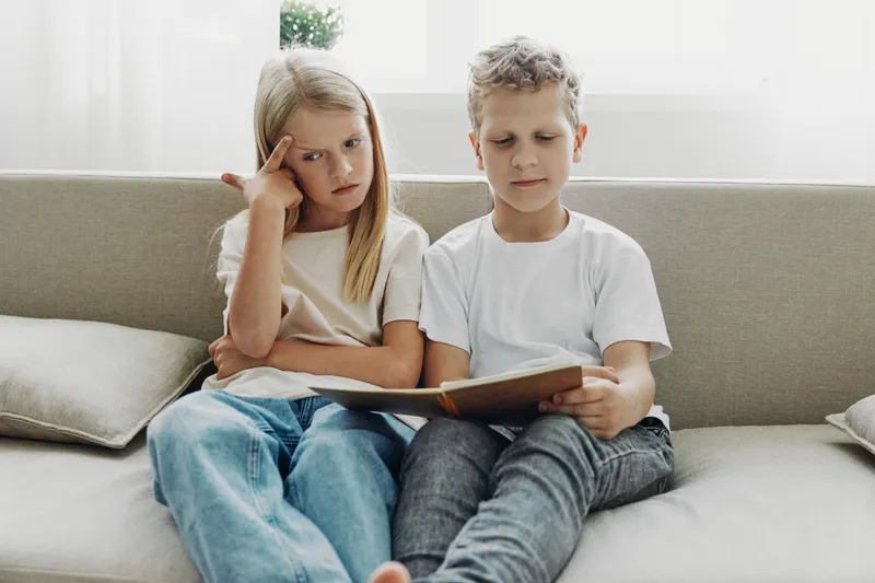 Two children sitting on sofa reading a book together, girl looks thoughtful while boy focuses on pages in bright living room setting.