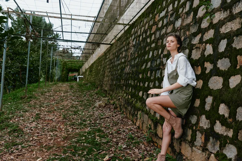 Young model in casual summer outfit poses gracefully against a rustic stone wall in a serene garden setting, showcasing a relaxed and confident vibe with natural warmth