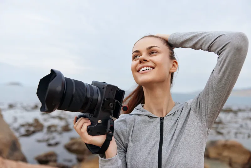 Happy young woman holding a camera on a rocky shore, enjoying nature photography, showcasing a vibrant outdoor lifestyle and capturing beautiful moments