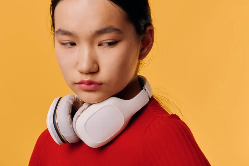 Young woman wearing white headphones around neck and red sweater standing against orange background with thoughtful expression and looking sideways in studio portrait.