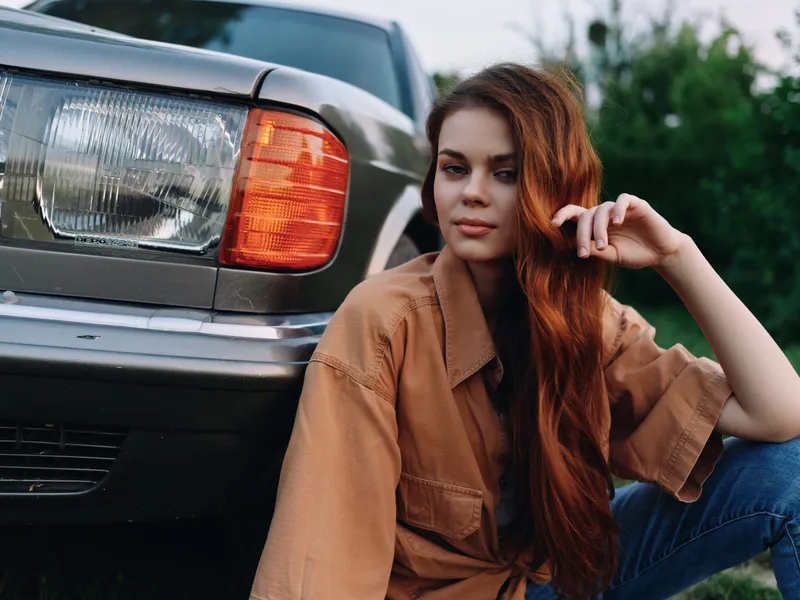 Young woman with long red hair in oversized shirt posing beside a vintage car in a green outdoor setting, evoking a relaxed and carefree vibe.
