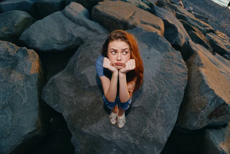 thoughtful woman sitting on large rocks with casual clothes and red hair outdoors, expressing pensive mood during daytime
