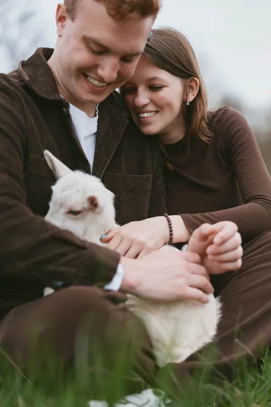 Young couple sitting on grass lovingly holding a white baby goat while smiling and enjoying a relaxed moment outdoors in casual brown clothing during a lifestyle photoshoot.
