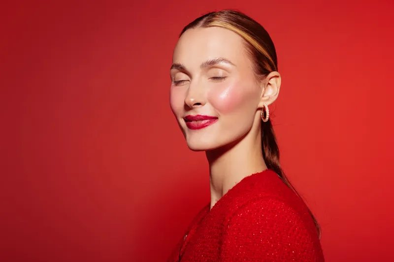 Portrait of a smiling woman with closed eyes wearing red lipstick and red sweater against red background. Concept of beauty, makeup, fashion, and confidence.