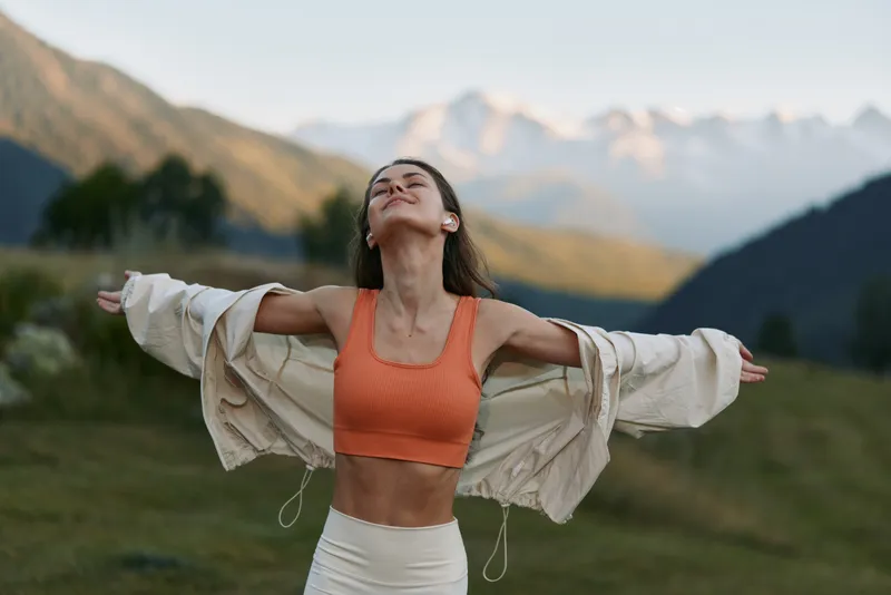 A carefree woman stands outdoors with arms spread wide, wearing an orange sports bra and a light robe. She breathes fresh air and savors freedom amid majestic mountain scenery, inspiring vitality.