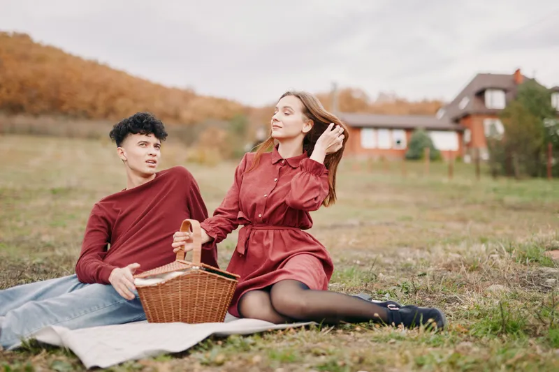 A young couple enjoying a candid autumn picnic on a rustic hillside, sharing a woven basket and relaxed conversation with an authentic, genuine atmosphere of everyday life and simple seasonal rituals.