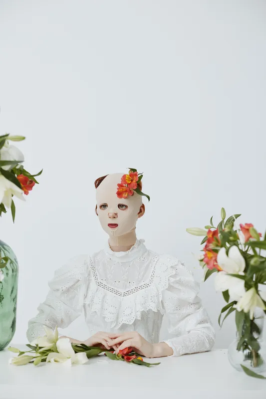 A conceptual still life featuring a mannequin with a floral headpiece, dressed in a vintage white dress, surrounded by blooming flowers in a minimalist white background