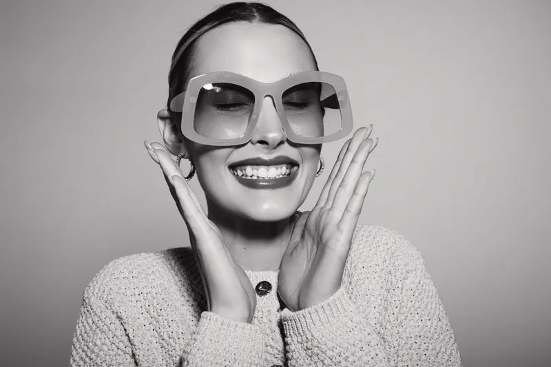 Smiling white woman with fair skin wearing large sunglasses and knitted sweater, posing with hands near face in studio light.