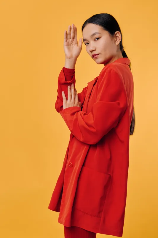Young woman in red coat and sweater posing with raised hand against orange background. Fashion model with long black hair, looking confident and stylish in studio portrait.