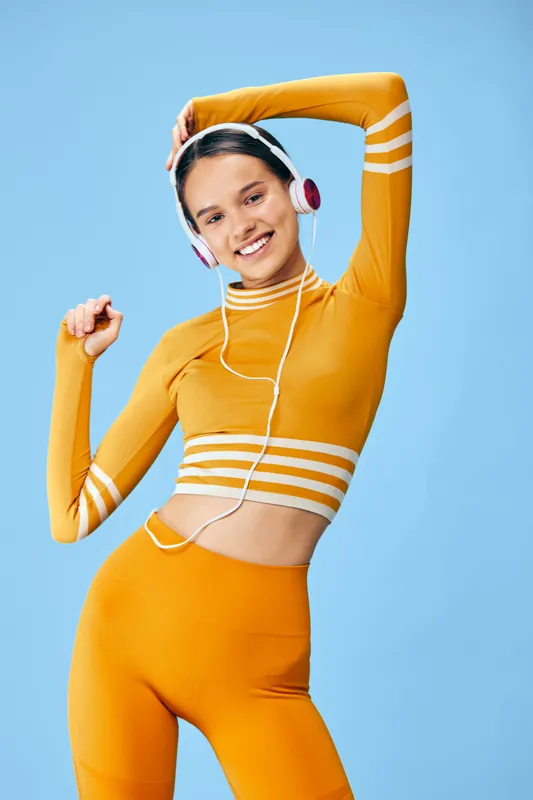 Woman fitness athlete in vibrant orange activewear stretching with headphones, smiling confidently against a clean light blue studio background, energetic sporty lifestyle concept.