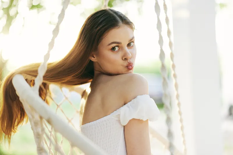 Young woman with long hair playfully posing on a swing, wearing an off shoulder white dress in a sunny garden setting, evoking feelings of joy and relaxation