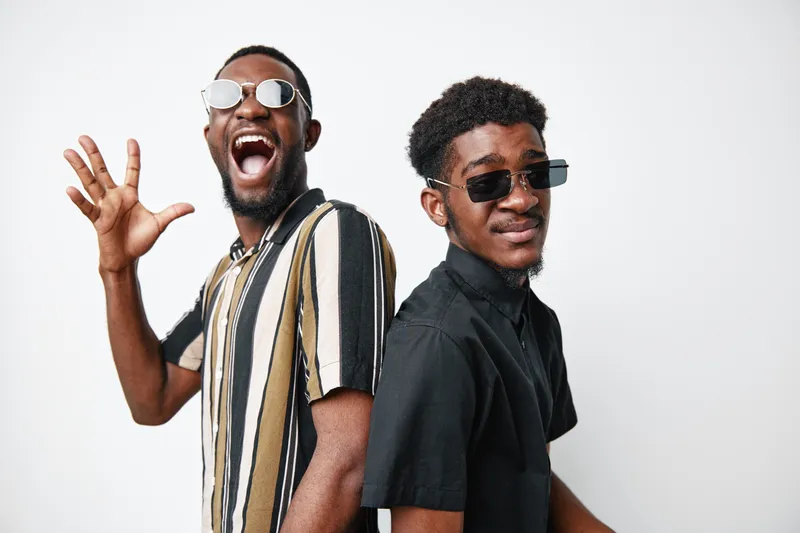 Two African American men with dark skin wearing sunglasses and casual shirts posing back to back on white background with confident and joyful expressions.