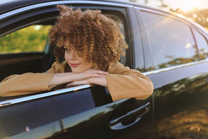 Smiling woman with curly hair leaning on a car window in natural light, casual lifestyle moment with outdoors vibe and retro film color, warm and relaxed