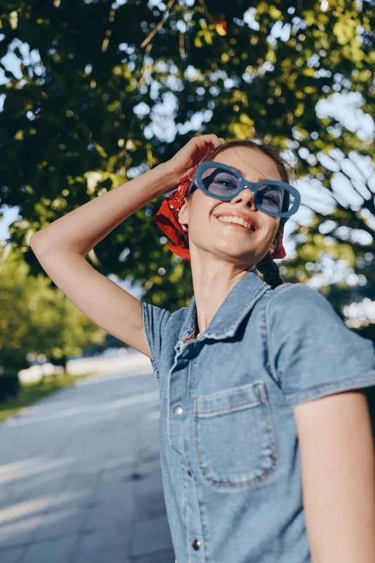 Lifestyle woman wearing modern boho-western denim and blue sunglasses, smiling outdoors with natural light and green background, film color effect enhancing street style.