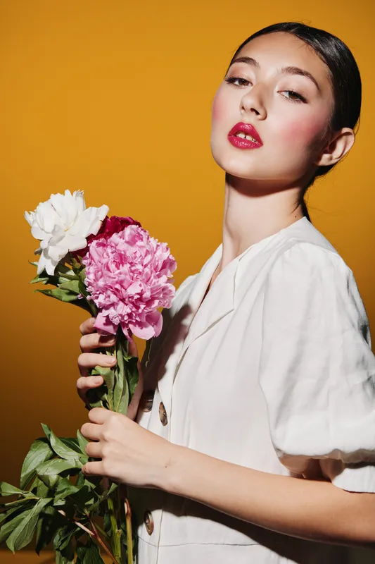 Young fashionable woman with bright makeup poses with pink and white flowers against an orange background. Concept of beauty creative fashion and floral style.