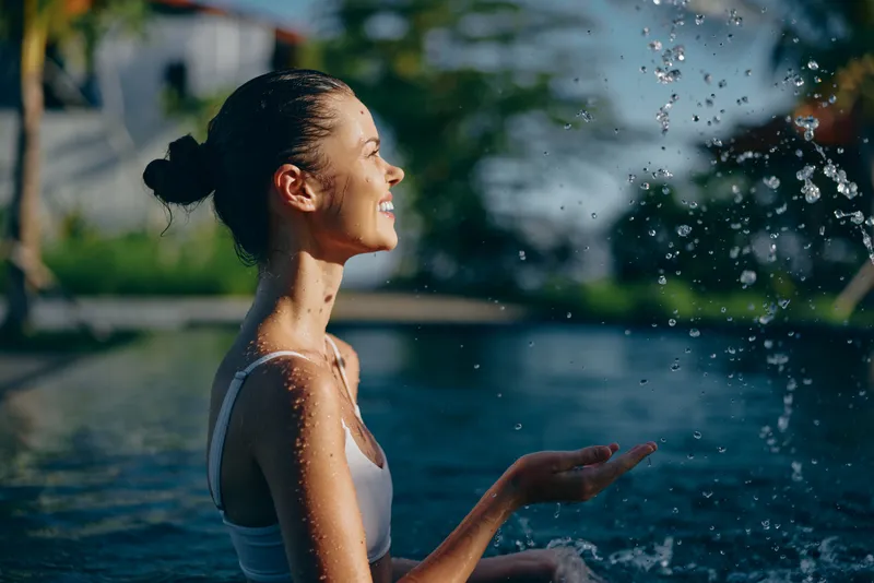 Young woman enjoys outdoor pool water splashing on her face with closed eyes and smiling expression, feeling relaxed lifestyle moment in bright sunny day.