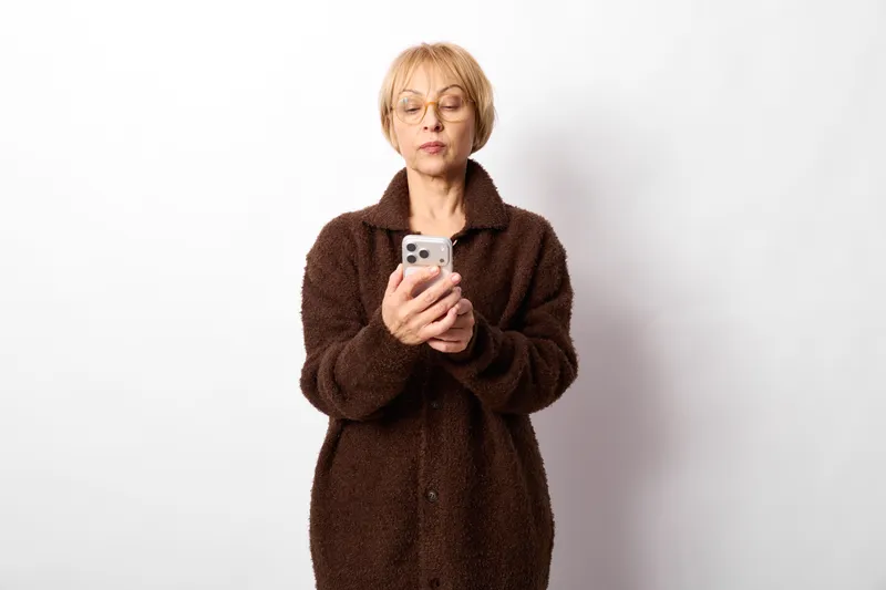 Woman smartphone selfie portrait of a senior female standing on white studio background, calm expression, casual coat, looking at screen, copy space.