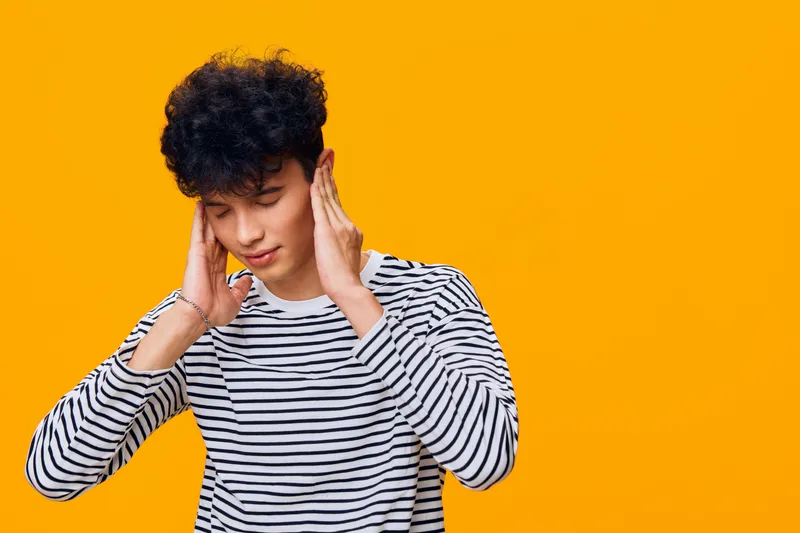young man covering ears with hands, curly hair, striped shirt, frustrated expression, bright orange background, emotional discomfort