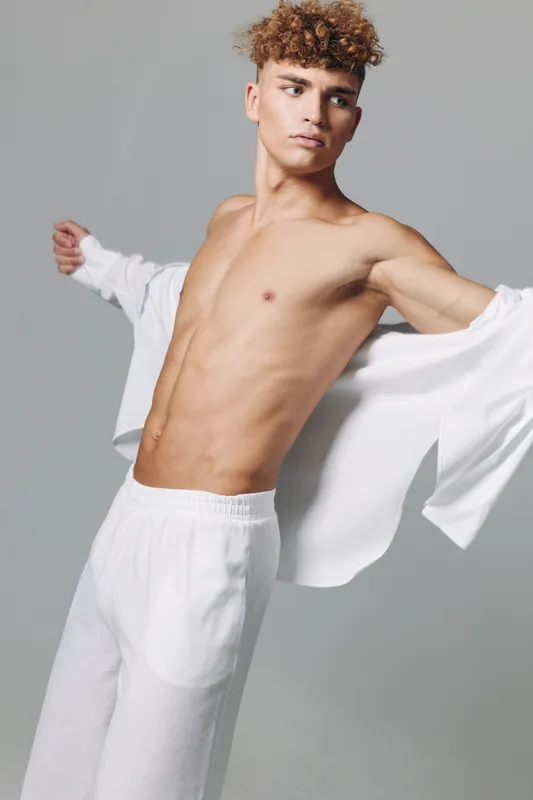 Young man with curly hair wearing white pants and removing white shirt, posing against gray background with focused expression and toned body in studio.