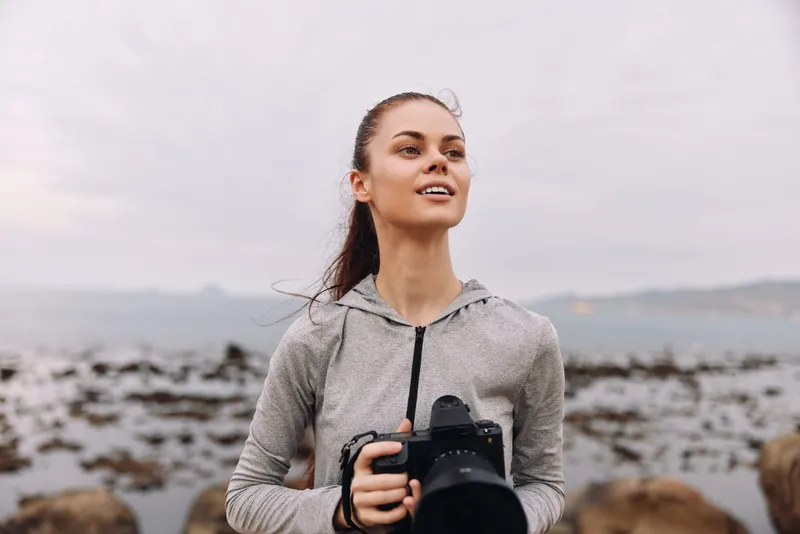 Young woman with a camera at the beach, smiling joyfully while embracing her passion for photography, wearing a gray sports hoodie in a coastal setting