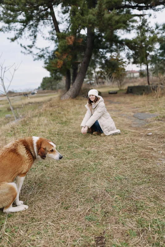 Outdoor scene with a woman kneeling on the grass in a park, talking on a phone while a calm dog sits nearby, capturing peaceful companionship and everyday outdoor moments amid trees