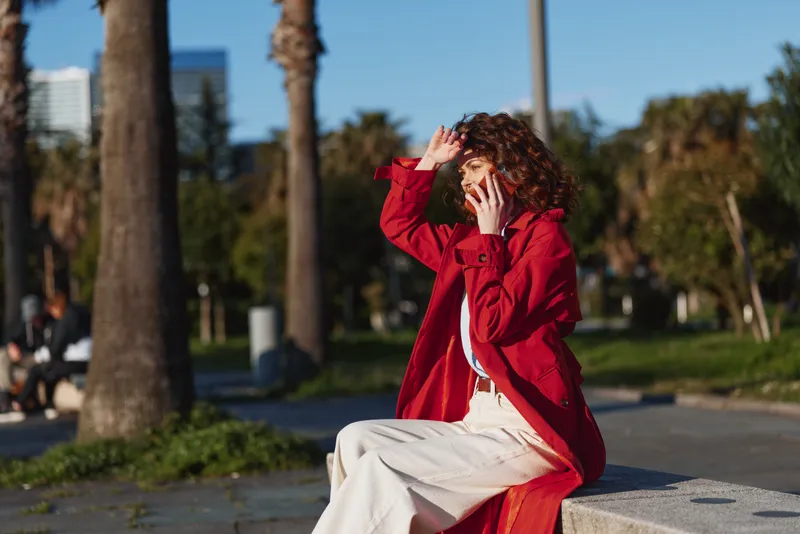 A young woman with curly hair sits on the pavement in a red coat, shielding her eyes from bright sun in a sunny park, conveying a candid moment outdoors.