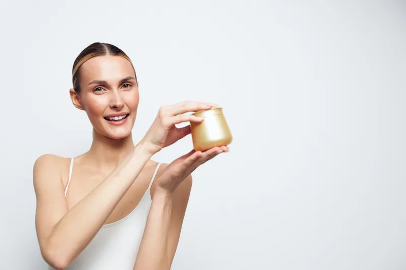 Smiling woman, skincare routine, holding a face cream jar near her cheek, close up on clean white background, wellness and self care concept, soft studio lighting, calm