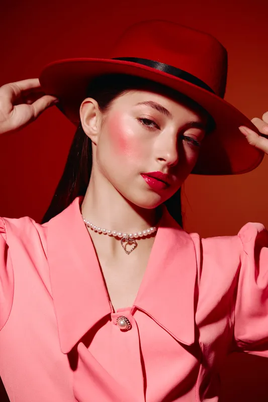 Young female beauty model in vibrant red hat and pink outfit poses against red background. Glossy skin and bold lips emphasize fashion and creative makeup concept.