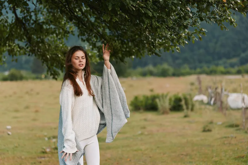 A woman in a cozy knit sweater stands beneath a leafy tree in an open field, holding a soft blanket over her arm. Bright natural light highlights a calm, casual lifestyle moment outdoors.