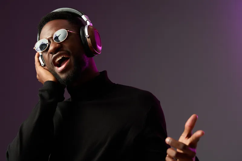 Energetic young Black man enjoying music with headphones, wearing sunglasses, against a gradient background, conveying joy and passion for music