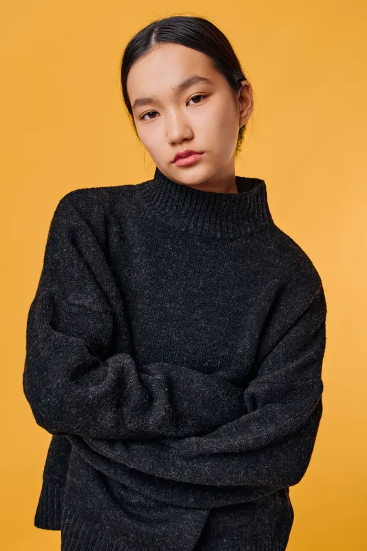 Young woman with dark hair wearing a black sweater poses with folded arms against orange background. Portrait of calm fashion model with natural makeup and casual style for winter season.