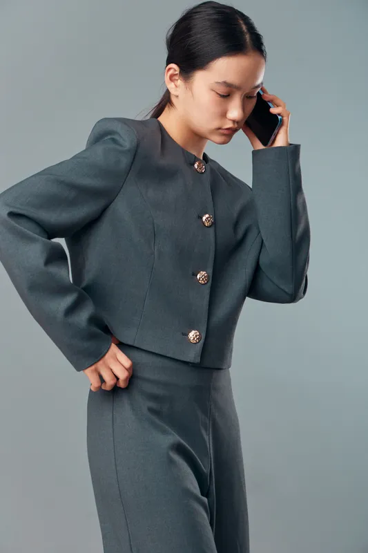 Young woman in grey pantsuit talking on phone, standing with one hand on hip and serious expression against plain background for communication and business.