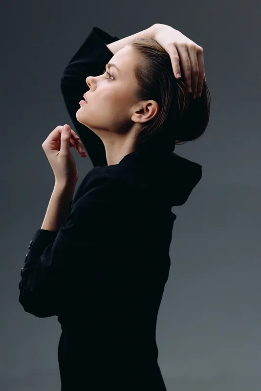 Profile of thoughtful woman in black clothing posing against gray background with minimalistic style and soft lighting
