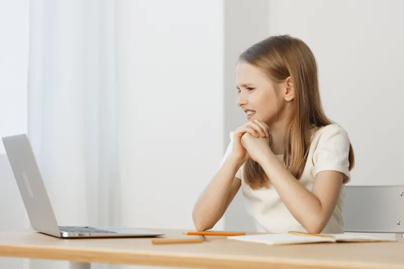 Young girl in a light colored dress, smiling at a laptop, sitting at a wooden desk with stationery, expressing happiness and concentration in a bright, minimalistic room