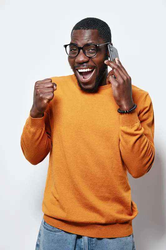 African American man with dark skin wearing glasses and orange sweater talking on phone and celebrating success with fist pump on white background.