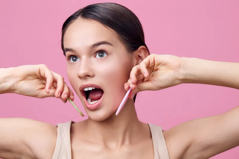 Young woman expressing joy while holding colorful hair removal wax sticks against a pink background, showcasing beauty and skincare concepts
