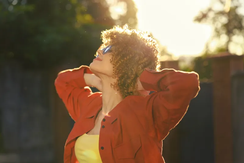 Smiling woman with curly hair outdoors, backlit by sun, wearing an orange jacket and sunglasses. Casual lifestyle vibe, warm glow, and confident stance convey happiness and relaxation